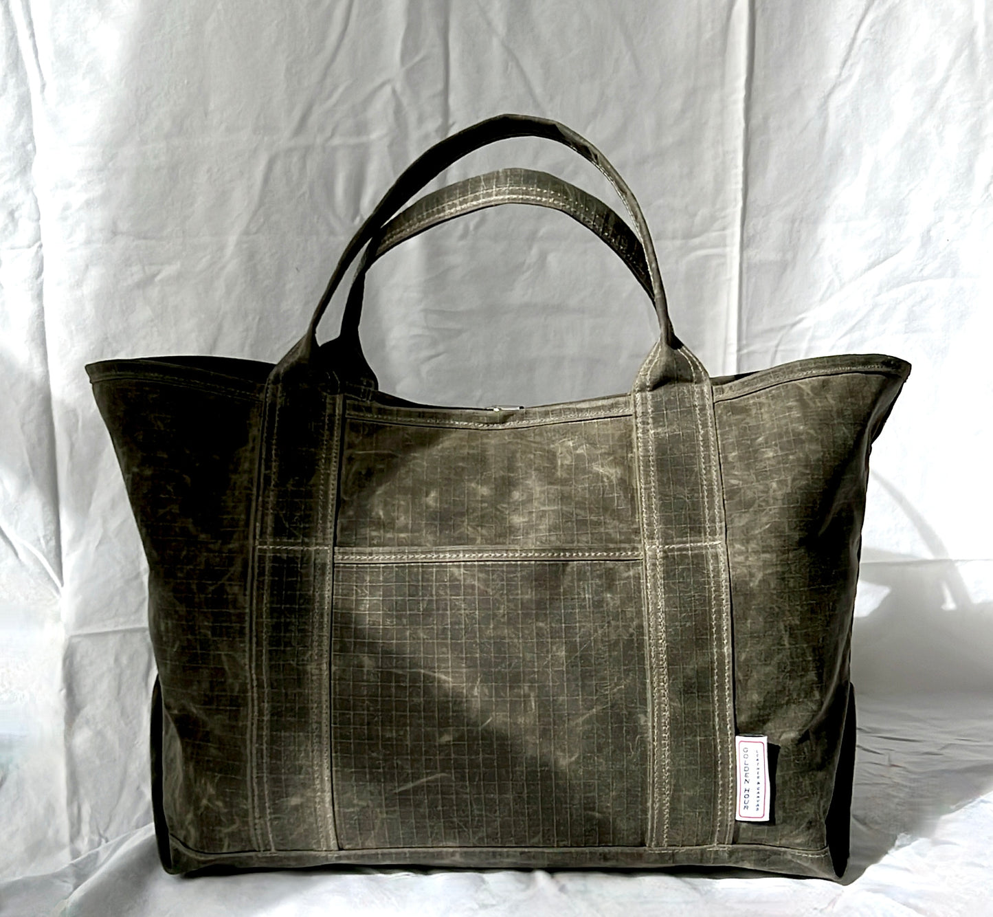 A large dark olive green canvas tote bag with a distressed, washed texture, featuring dual top handles, a front pocket, and a small white brand tag on the lower right side, set against a white fabric backdrop.